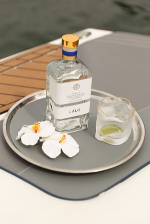 Clear tequila bottle on a silver serving tray aboard a boat, paired with a glass of ice and lime and two white hibiscus flowers on a teak deck with water in the background