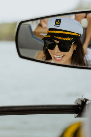 Reflection of a smiling woman in a captain’s hat and dark sunglasses captured in a boat mirror during a sunny day on calm water.