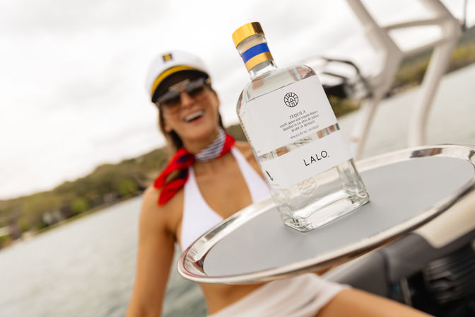 Clear tequila bottle on a silver tray aboard a motorboat, with a smiling woman in a captain's hat, sunglasses and red neck scarf in soft focus against a lake shoreline — playful summer boating scene.