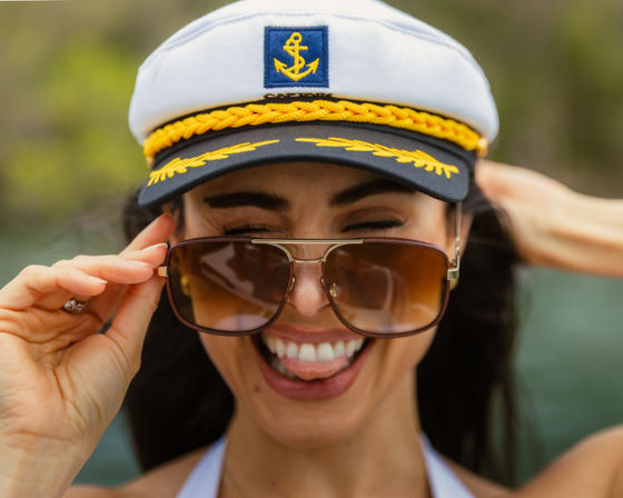 Playful woman on a boat wearing a white captain’s hat with a yellow anchor emblem and aviator sunglasses, smiling and sticking out her tongue against a blurred water and greenery background.