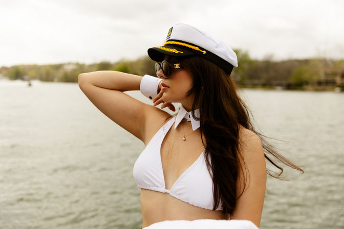 Stylish woman in a white captain’s hat and bikini top with sunglasses striking a playful pose on a boat with a lake and tree-lined shore in the background.