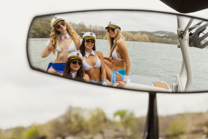 Reflection in a boat mirror of a playful group of four friends in captain hats, sunglasses and white bikinis smiling aboard a boat on a lake with a tree-lined shore.