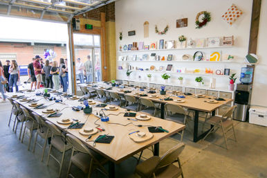 Open-garage craft studio set for a DIY workshop — long communal tables with wood rounds, hand tools and cords, wall shelves of artwork and plants, water cooler, and a small group gathered outside.