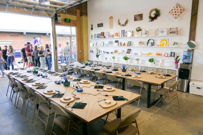Open-garage craft studio set for a DIY workshop — long communal tables with wood rounds, hand tools and cords, wall shelves of artwork and plants, water cooler, and a small group gathered outside.