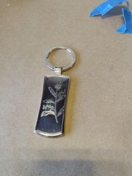 Silver keyring with a rectangular black pendant engraved with a delicate botanical sprig, photographed on kraft paper with a scrap of blue painter's tape
