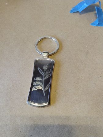 Silver keyring with a rectangular black pendant engraved with a delicate botanical sprig, photographed on kraft paper with a scrap of blue painter's tape
