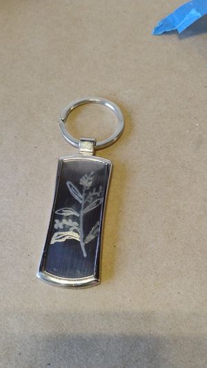 Silver keyring with a rectangular black pendant engraved with a delicate botanical sprig, photographed on kraft paper with a scrap of blue painter's tape