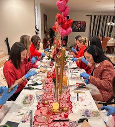 Indoor Valentine's DIY craft night: a group of women in red wearing blue gloves make floral resin trays at a long dining table decorated with heart balloons, candles, and flowers.