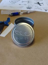 Small round metal tin with an etched lid showing three connected hearts and the date "4-30-22" on a brown craft-paper workspace, surrounded by a pen, paper stencils and blue painter's tape.