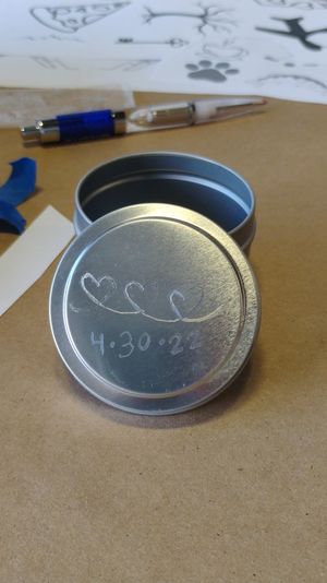 Small round metal tin with an etched lid showing three connected hearts and the date "4-30-22" on a brown craft-paper workspace, surrounded by a pen, paper stencils and blue painter's tape.
