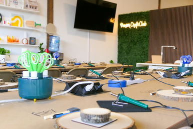 Indoor craft workshop table set for a DIY class with soldering irons, green scissors caddy, wood-slice workpieces, power strips and tools, TV and neon wall accent in the background