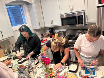 Three people doing a hands-on DIY craft party at a modern white home kitchen island with stainless-steel appliances, colorful art supplies, fresh flowers, snacks and drinks.