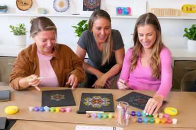 Three smiling women in a bright indoor art studio painting colorful dot-mandala designs on black canvases at a table lined with small paint pots, brushes and potted plants — DIY craft workshop scene