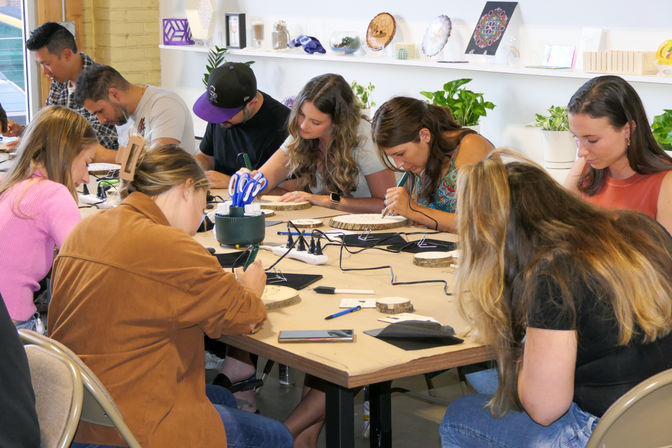 Adults gathered around a large table in a community art studio, focused on woodburning and decorating round wood slices with plants and artwork on shelves in the background