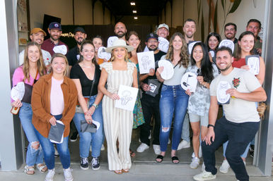 About 20 smiling adults pose at a studio entrance after a casual art workshop, many holding hand-drawn sketches (mushrooms, leaves, owl); woman in a striped jumpsuit and wide-brim hat stands front and center.