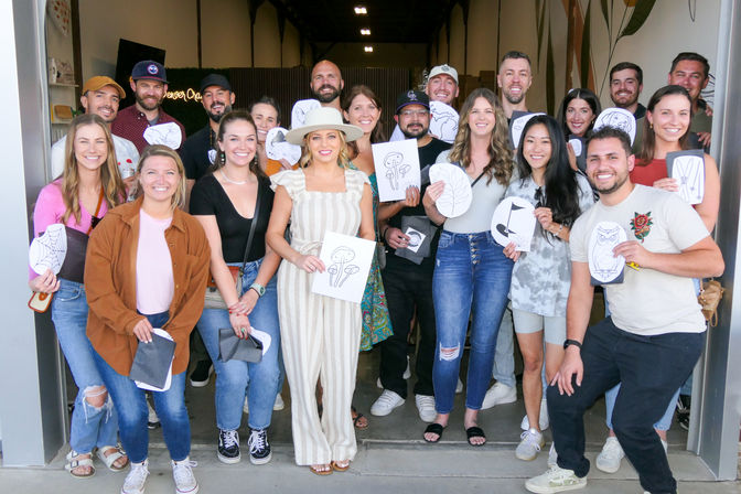 About 20 smiling adults pose at a studio entrance after a casual art workshop, many holding hand-drawn sketches (mushrooms, leaves, owl); woman in a striped jumpsuit and wide-brim hat stands front and center.