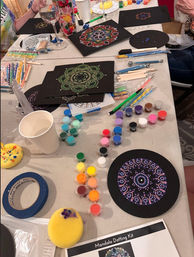 Indoor craft table with mandala dot painting kit: black circular canvases and panels featuring colorful dotted mandala designs, rows of small paint pots, dotting tools and brushes, sponges, masking tape and a paper cup for a DIY art session.