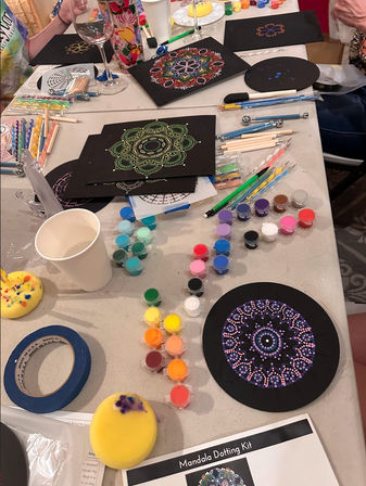 Indoor craft table with mandala dot painting kit: black circular canvases and panels featuring colorful dotted mandala designs, rows of small paint pots, dotting tools and brushes, sponges, masking tape and a paper cup for a DIY art session.