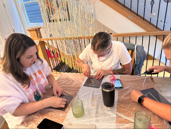 DIY craft night at a home dining table: three people shaping wire on black mats with scissors and pliers, tumblers and phones nearby, sparkly fringe backdrop and staircase.