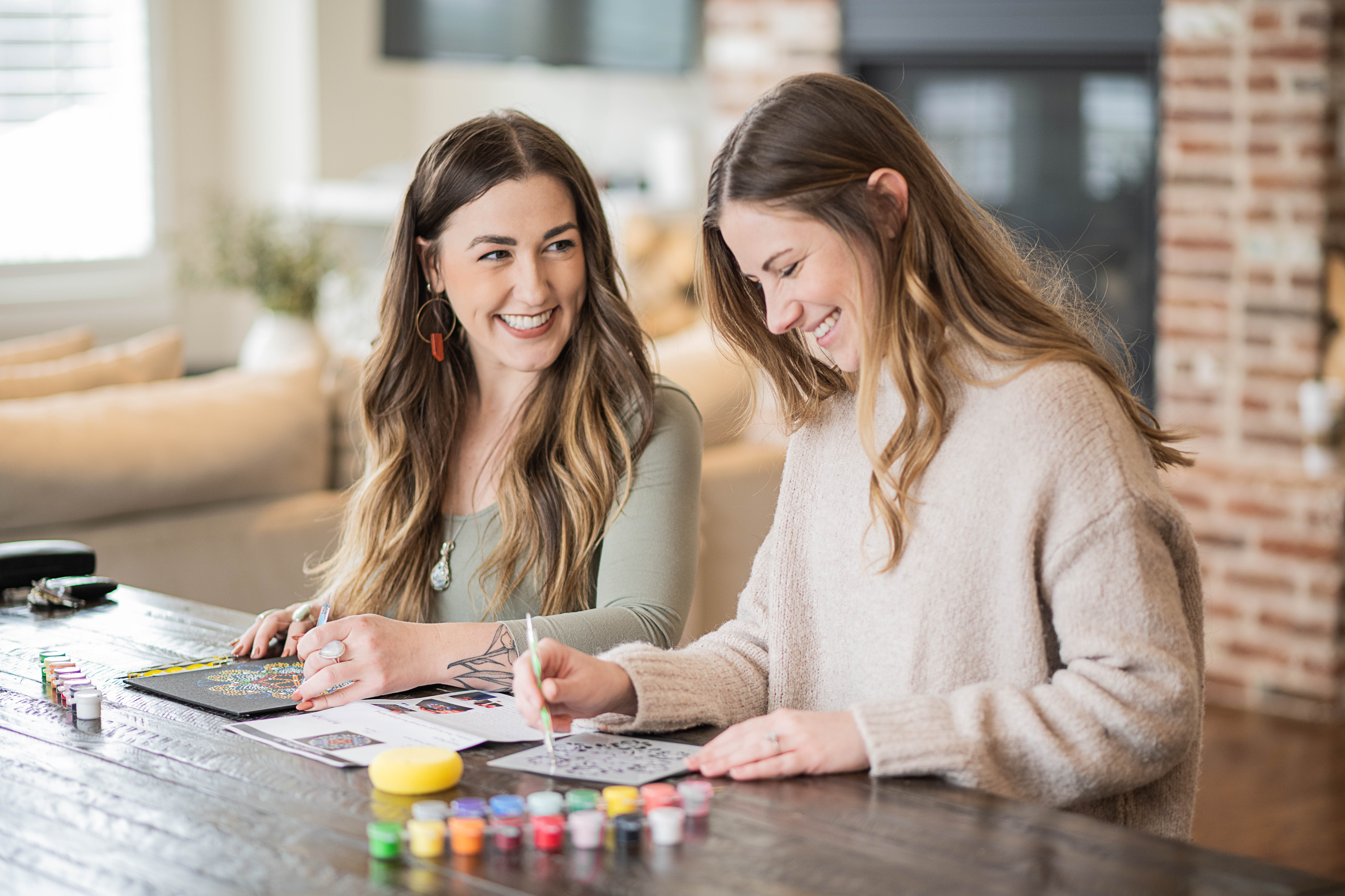 Two women laughing while painting colorful craft pages with small paint pots and brushes on a rustic wooden table in a bright, cozy living room
