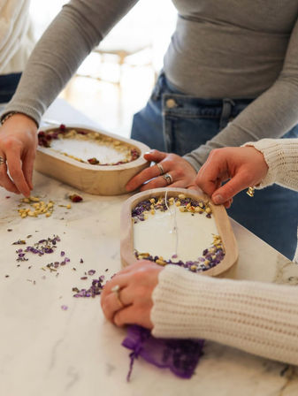Hands-on DIY candle making: creamy white candles poured in wooden oval molds garnished with dried purple and yellow flowers on a marble countertop.