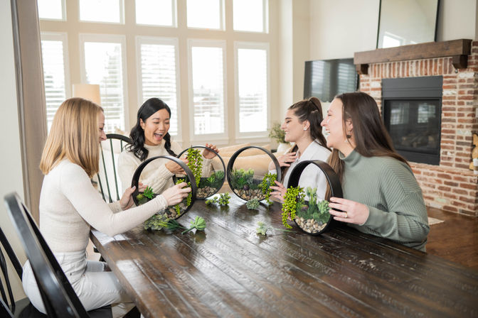 Four friends at a wooden dining table in a bright living room making circular succulent terrariums, laughing and arranging greenery near a brick fireplace.