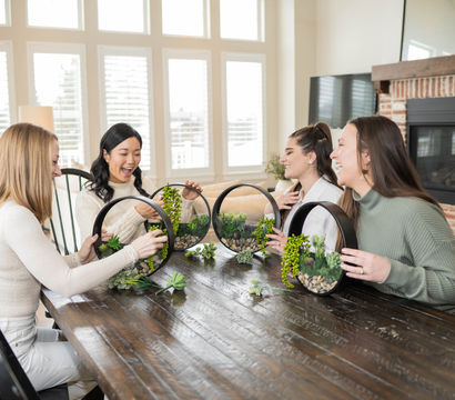 Four friends at a wooden dining table in a bright living room making circular succulent terrariums, laughing and arranging greenery near a brick fireplace.
