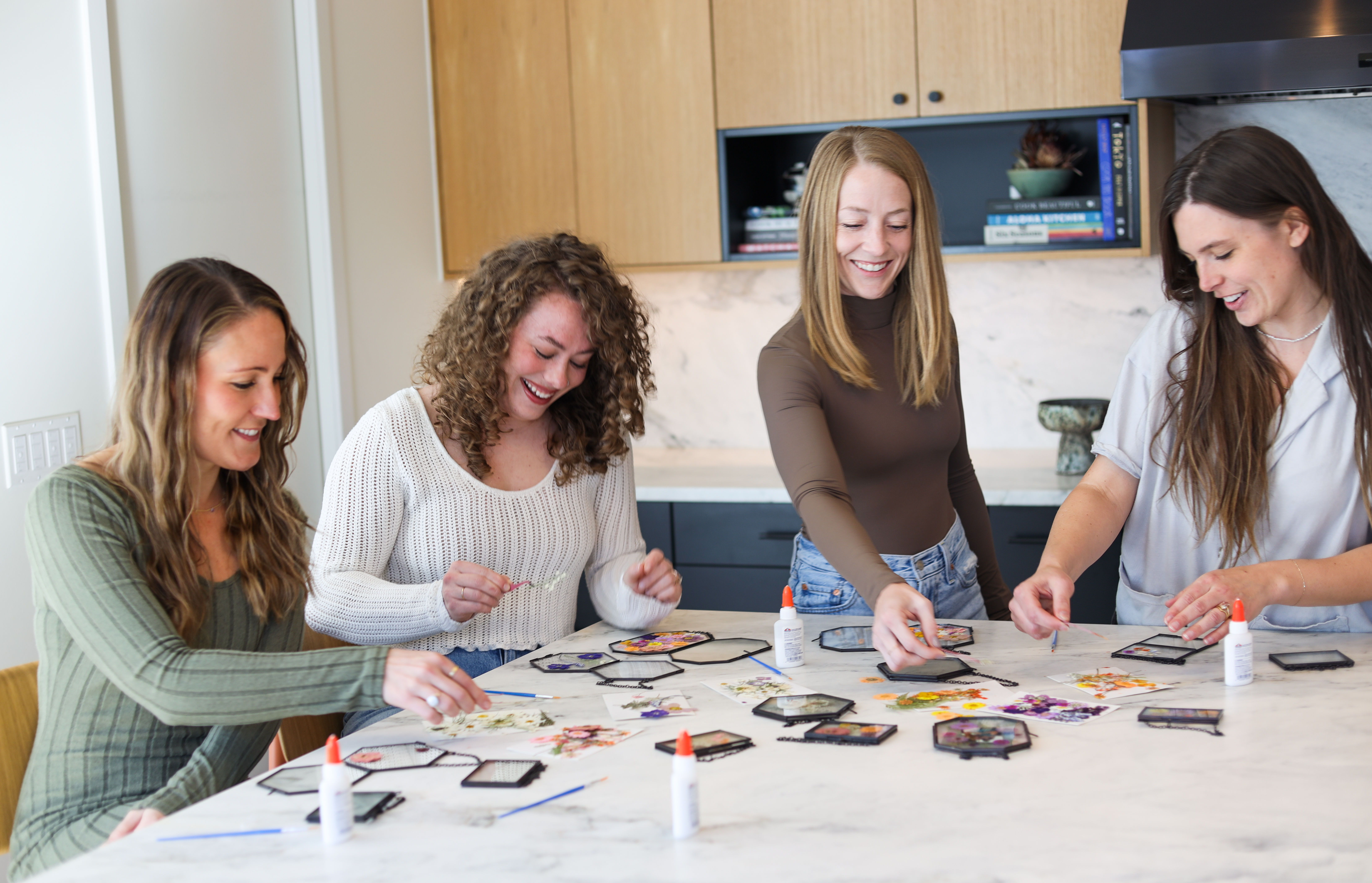 Four friends smiling around a marble kitchen island making DIY pressed-flower hexagon frames with glue