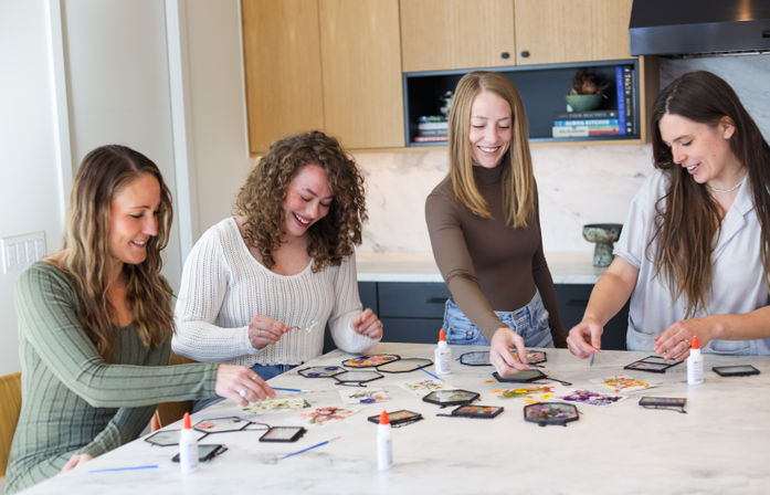 Four friends smiling around a marble kitchen island making DIY pressed-flower hexagon frames with glue