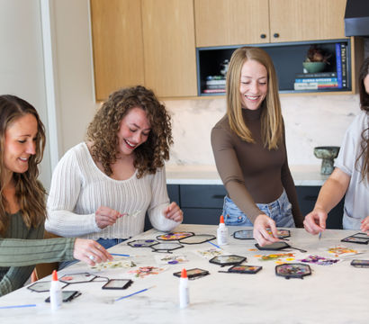 Four friends smiling around a marble kitchen island making DIY pressed-flower hexagon frames with glue