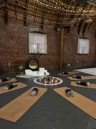 Cozy brick-walled loft studio set for a sound-healing yoga session with cork mats and rolled blankets arranged in a circle around candles, flowers, crystal singing bowls and a gong beneath twinkling string lights.