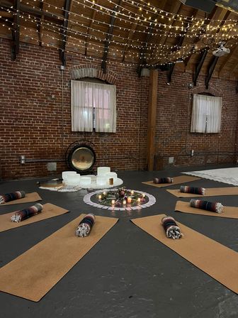 Cozy brick-walled loft studio set for a sound-healing yoga session with cork mats and rolled blankets arranged in a circle around candles, flowers, crystal singing bowls and a gong beneath twinkling string lights.