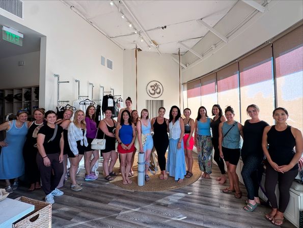 Sunlit community yoga studio with Om emblem, about 20 women in activewear gathered around aerial yoga straps and mats on a wooden floor