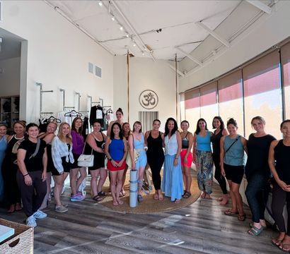 Sunlit community yoga studio with Om emblem, about 20 women in activewear gathered around aerial yoga straps and mats on a wooden floor