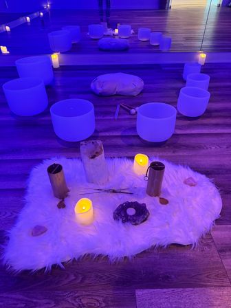 Blue-lit wellness studio set for a sound bath: circle of frosted crystal singing bowls and mallets around a meditation cushion reflected in a mirror, with a white faux-fur altar rug holding LED candles, crystals and tuning tubes on a wood floor.