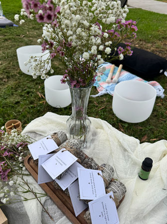 Outdoor wellness spread on a grassy park lawn: glass vase of pink and white wildflowers, tied sage smudge bundles with affirmation tags on a wooden tray, crystal singing bowls with mallets on a patterned blanket, and a small essential oil bottle.