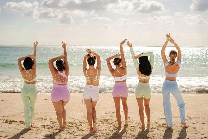 Six women in pastel activewear stand barefoot on a sunlit sandy beach facing a turquoise ocean, stretching their arms upward in a relaxed group yoga pose with sparkling water and soft clouds.
