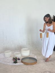 Woman in a white sundress, barefoot, holding wooden mallets over frosted crystal singing bowls on a white shag rug — minimalist sound-bath setup with flowers and a wooden tray in a bright studio.