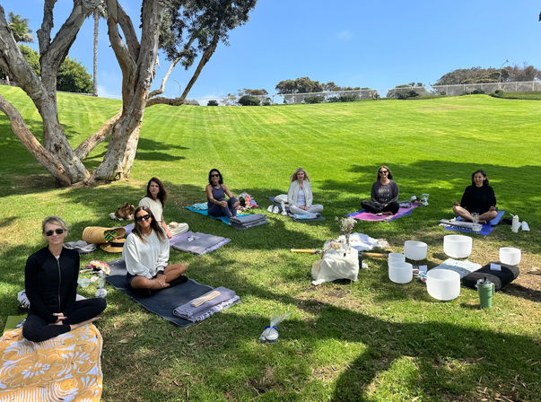 Group meditation and sound-bath session in a sunny park: participants seated on yoga mats and blankets on a grassy hillside under a large tree with crystal singing bowls arranged nearby.