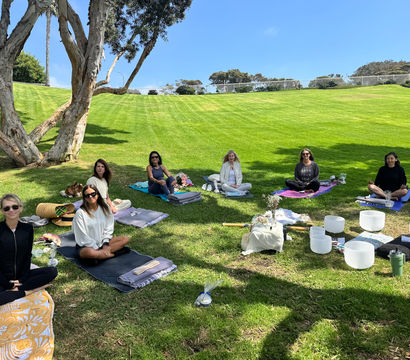 Group meditation and sound-bath session in a sunny park: participants seated on yoga mats and blankets on a grassy hillside under a large tree with crystal singing bowls arranged nearby.