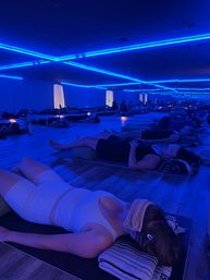 Soothing blue-lit yoga studio with a group in savasana lying on mats, eye towels and bolsters, mirrored wall reflecting neon LED lines