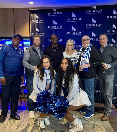 Smiling group photo in a blue-themed sports lounge — two white-and-blue cheerleaders kneel with shiny blue pom-poms in front of six adults in casual fan gear, some holding drinks.