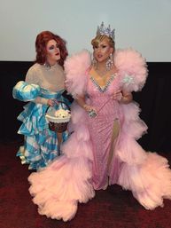 Two drag performers posing indoors at a show: one in a sparkly pink gown with voluminous pink tulle train, rhinestone crown, oversized jeweled earrings and star wand; the other in a blue-and-white ruffled dress holding a wicker basket with a plush toy.