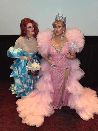 Two drag performers posing indoors at a show: one in a sparkly pink gown with voluminous pink tulle train, rhinestone crown, oversized jeweled earrings and star wand; the other in a blue-and-white ruffled dress holding a wicker basket with a plush toy.