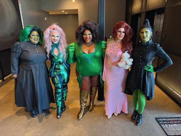 Group of five drag performers posing in a venue lobby, wearing dramatic wigs and makeup and vibrant costumes — two in green sequins and boots, one in a sparkling pink gown holding a teddy bear, and two in black outfits with green accents.