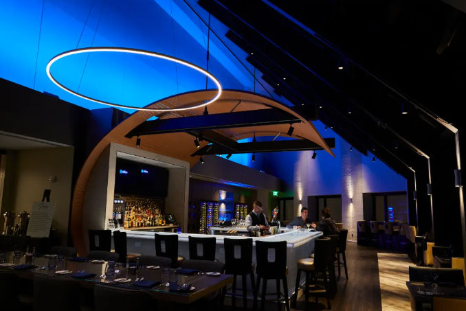 Upscale modern bar and lounge with vaulted ceiling washed in blue light, circular LED ring fixture above a marble-topped central bar lined with stools and bartenders serving guests.