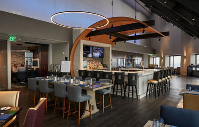 Upscale modern hotel bar and lounge interior with a central white marble bar, high stools, communal dining table, circular pendant light and arched wooden canopy over dark wood floors and large windows.