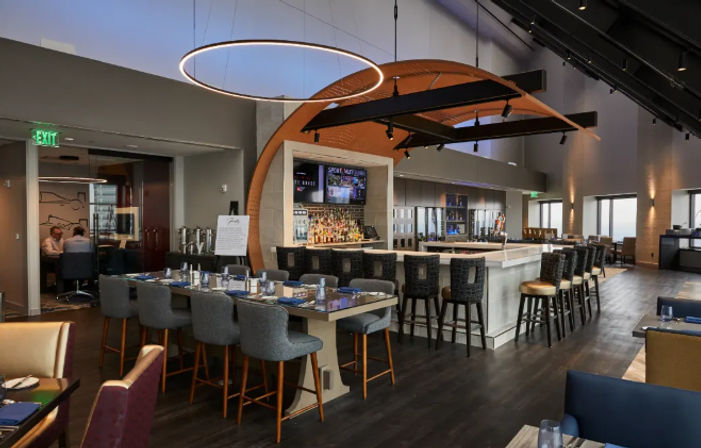 Upscale modern hotel bar and lounge interior with a central white marble bar, high stools, communal dining table, circular pendant light and arched wooden canopy over dark wood floors and large windows.