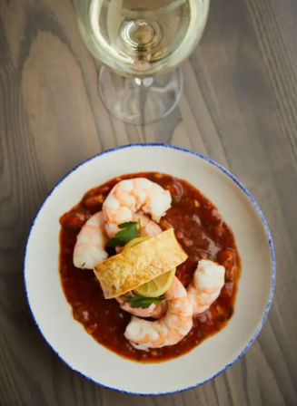 Plump shrimp cocktail seafood appetizer in zesty tomato-based sauce, lemon slice and parsley garnish with a crusty crostini and a glass of white wine on a rustic wooden table