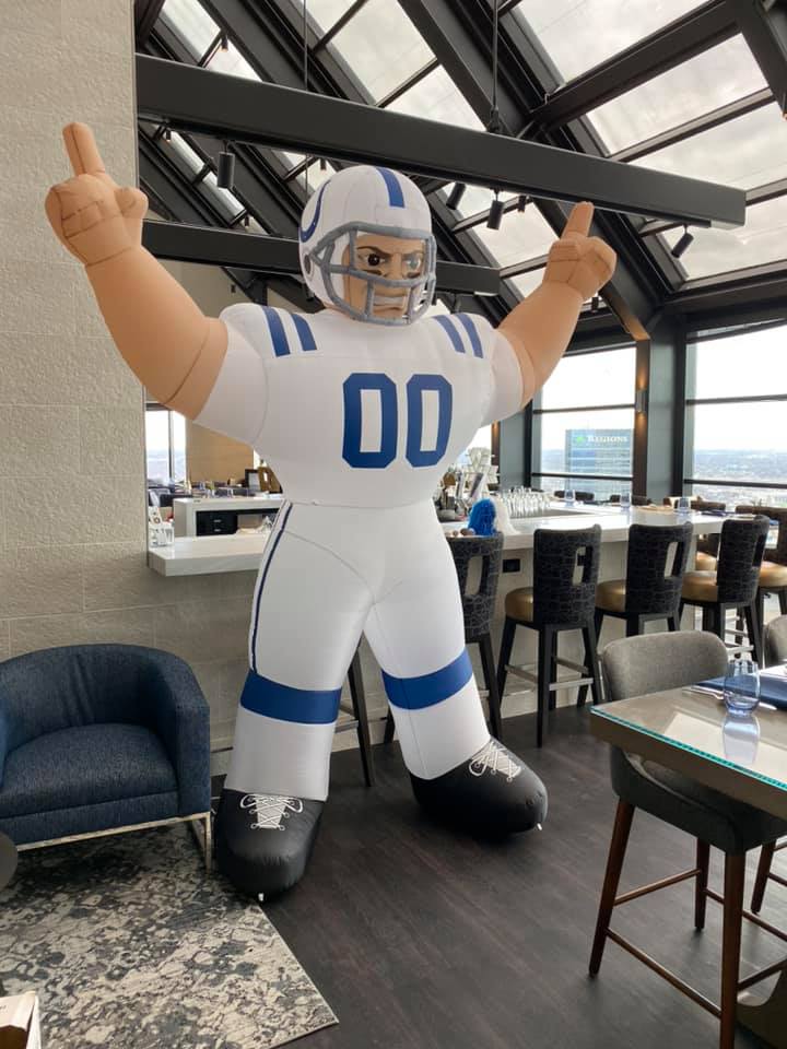 Inflatable giant football player mascot in white-and-blue uniform (00) inside a rooftop lounge with panoramic city skyline views